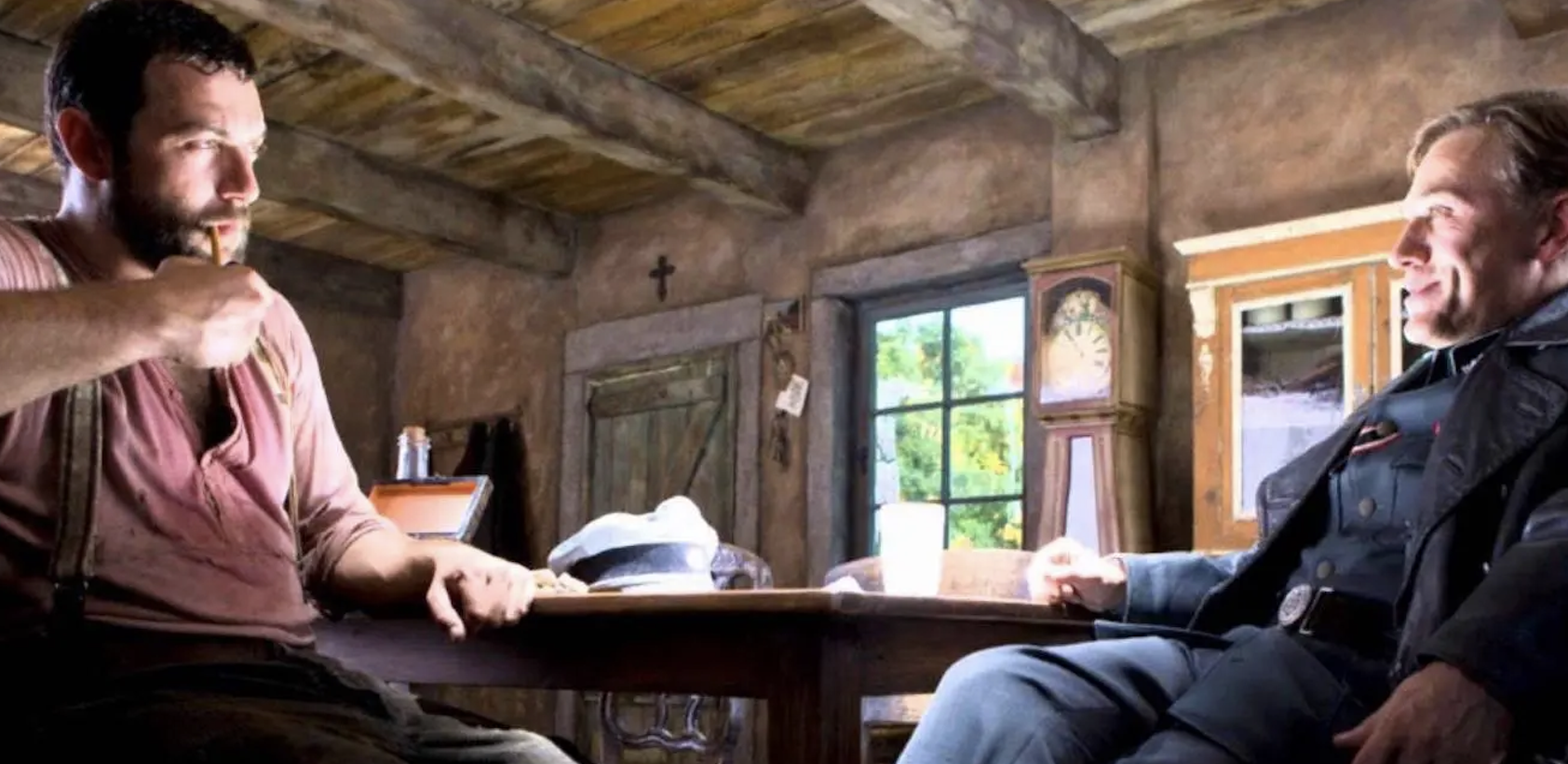 Two men, one in casual attire and the other in a uniform, sit at a wooden table in a rustic room lit by natural light