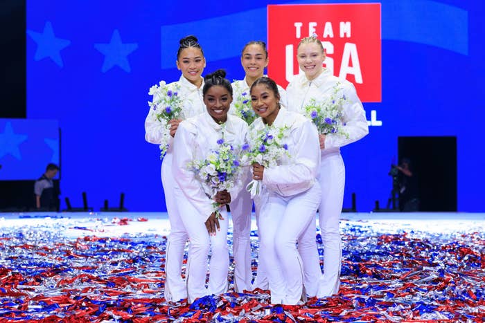 Simone Biles, Suni Lee, Jordan Chiles, Grace McCallum, and Jade Carey hold bouquets, posing together on a stage with confetti, in front of a &quot;Team USA&quot; background
