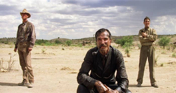Three men stand in a barren landscape, one kneeling in the foreground with oil on his face and a serious expression