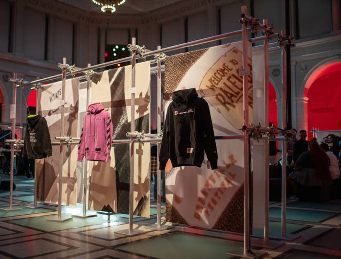 Fashion exhibit featuring multiple hoodies displayed on a metal framework, located in a stylish indoor setting with arched windows and red lighting accents