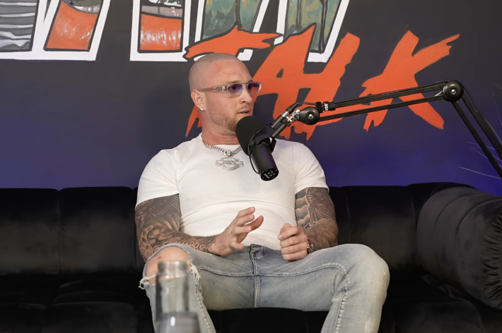A bald man with tattoos, wearing a white t-shirt and jeans, speaks into a microphone while seated on a black couch, on a podcast set with vibrant wall art