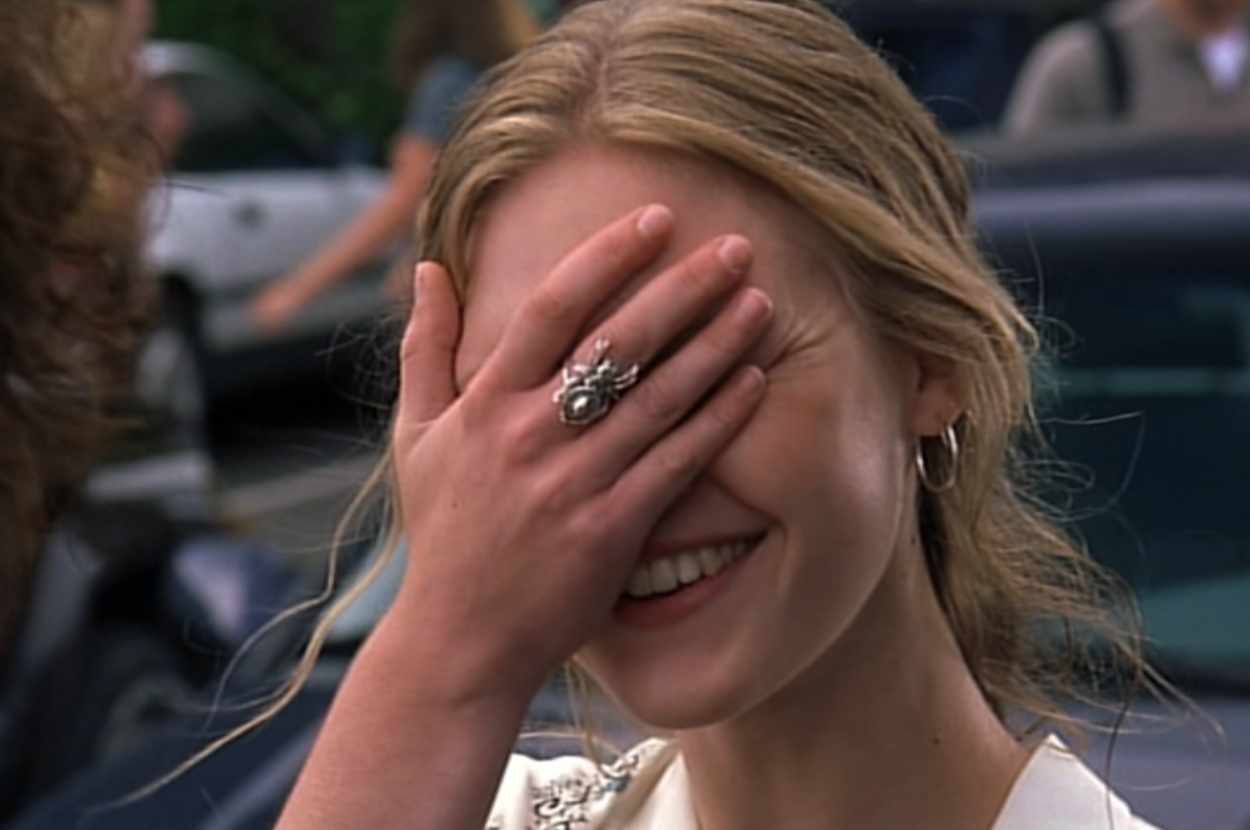 Woman smiling and covering her face with her hand, wearing a ring and an earring, outdoors with people and cars in the background