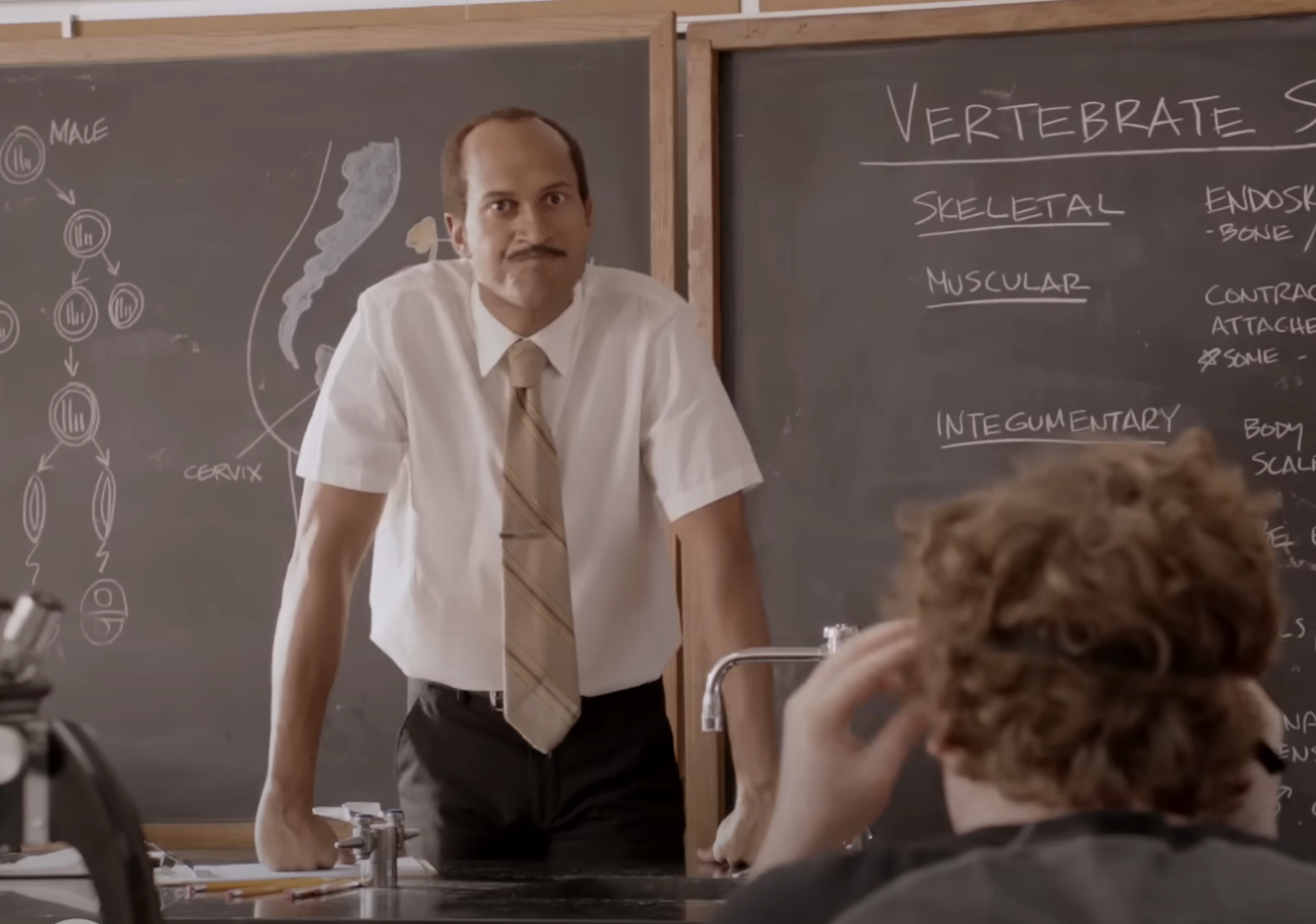 Keegan-Michael Key stands in a classroom leaning on a desk, teaching a lesson on vertebrate anatomy with detailed notes on a chalkboard in a scene from a comedy skit