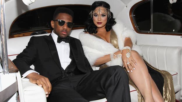 A man in a tuxedo and sunglasses sits with a woman in a gold gown and fur stole on a luxurious car's backseat