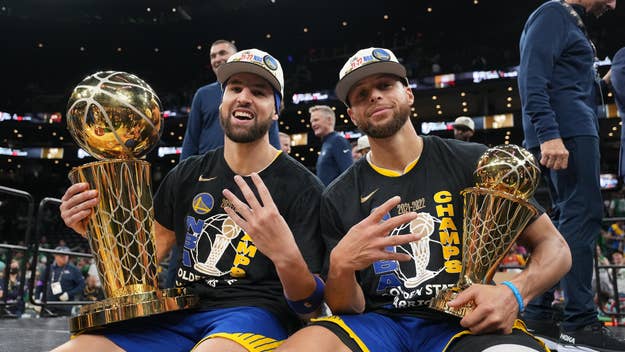 Klay Thompson and Stephen Curry sit holding NBA championship trophies while wearing "Champs" t-shirts and hats, showing four fingers to indicate their titles