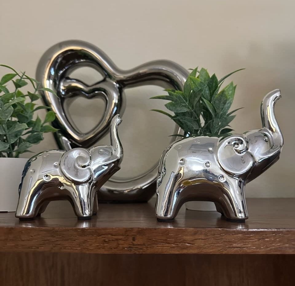 Two silver elephant figurines stand on a wooden shelf in front of green plants and a heart-shaped metallic sculpture