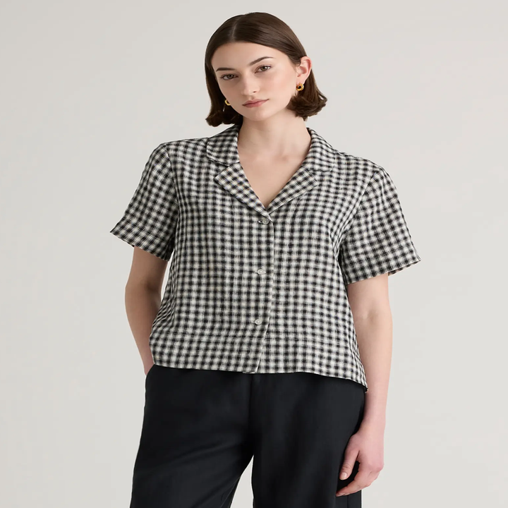 Model wearing a checkered short-sleeve blouse and black pants, posing against a neutral background