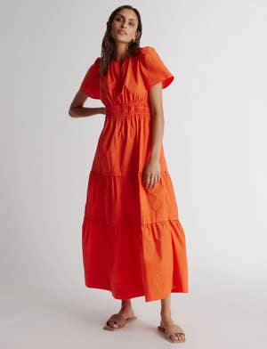 Model in an orange maxi dress with short sleeves and tiered skirt, standing with one hand on her hip, wearing sandals