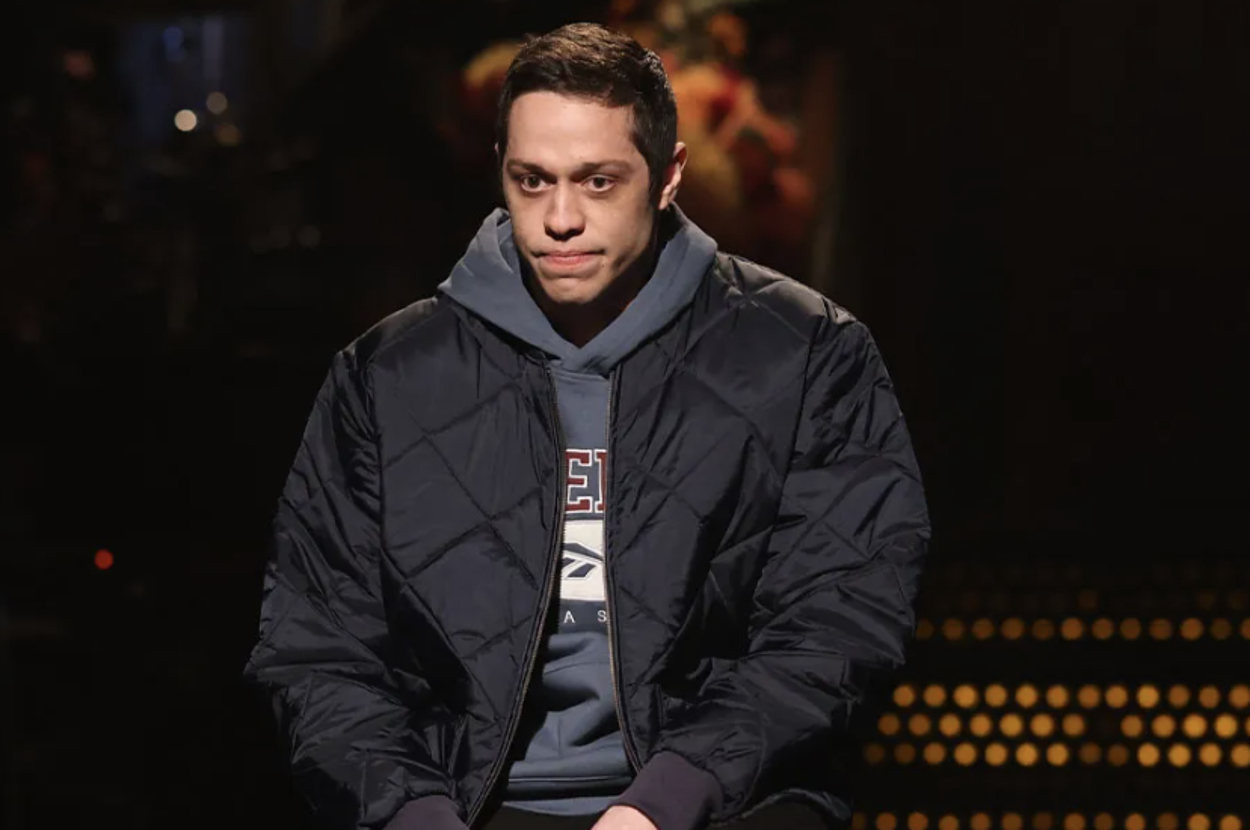 Pete Davidson sits on a dark set wearing a casual hoodie and a quilted jacket, looking introspective