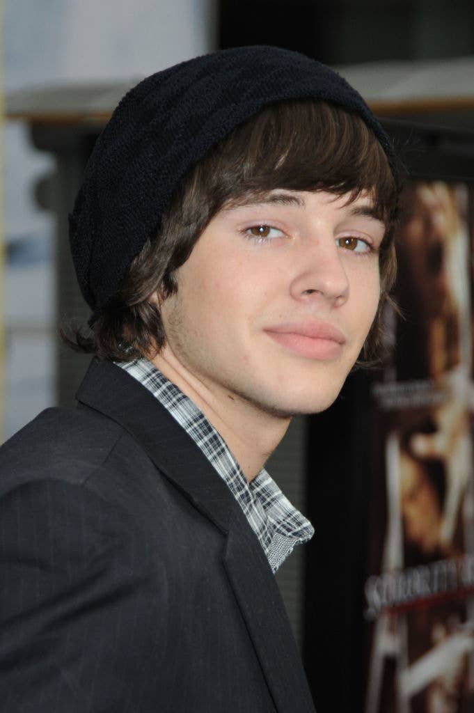 A young man with shoulder-length hair wearing a black beanie, plaid shirt, and blazer, standing indoors