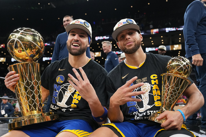 Klay Thompson and Stephen Curry seated, wearing "Champs" shirts and caps, each holding a trophy and showing 4 fingers