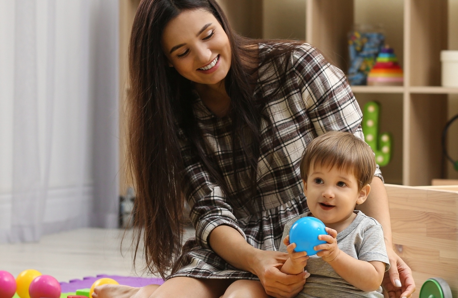 A woman in a plaid dress sits on the floor, playing with a toddler holding a blue ball, surrounded by colorful toys