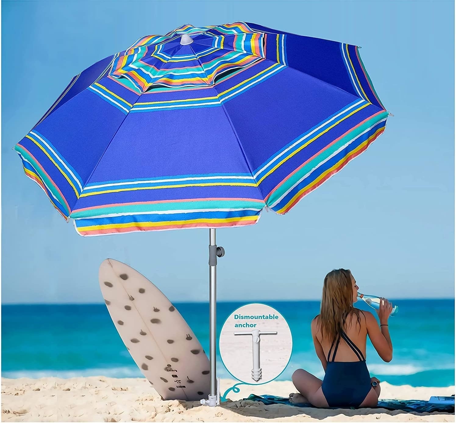 A model sits on a beach under a blue-striped umbrella near a surfboard