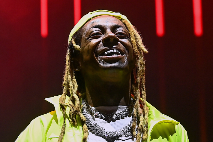 Lil Wayne, smiling broadly, stands under stage lights in a sporty outfit with layered necklaces and a cap, showing his distinctive dreadlocks