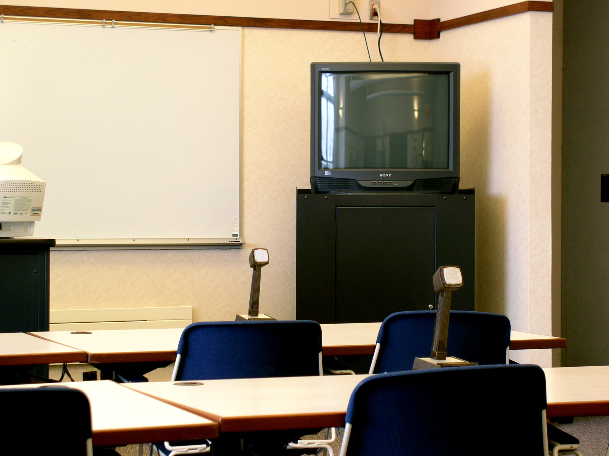 Empty classroom with several desks and chairs, a whiteboard on the wall, and a TV on a stand at the front of the room
