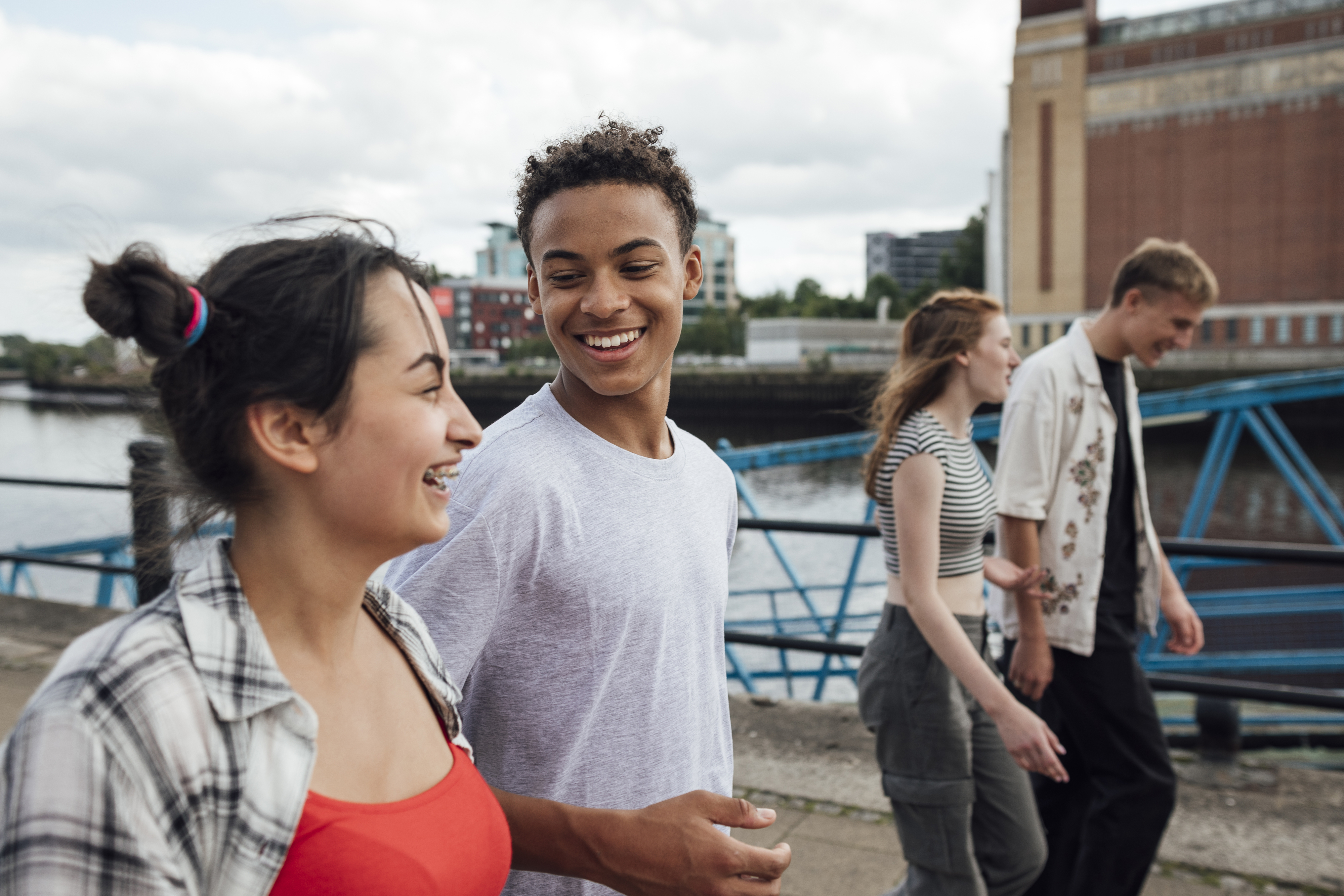 Two people are chatting and laughing while walking beside a waterfront. Two other people are walking in the background. Names unknown