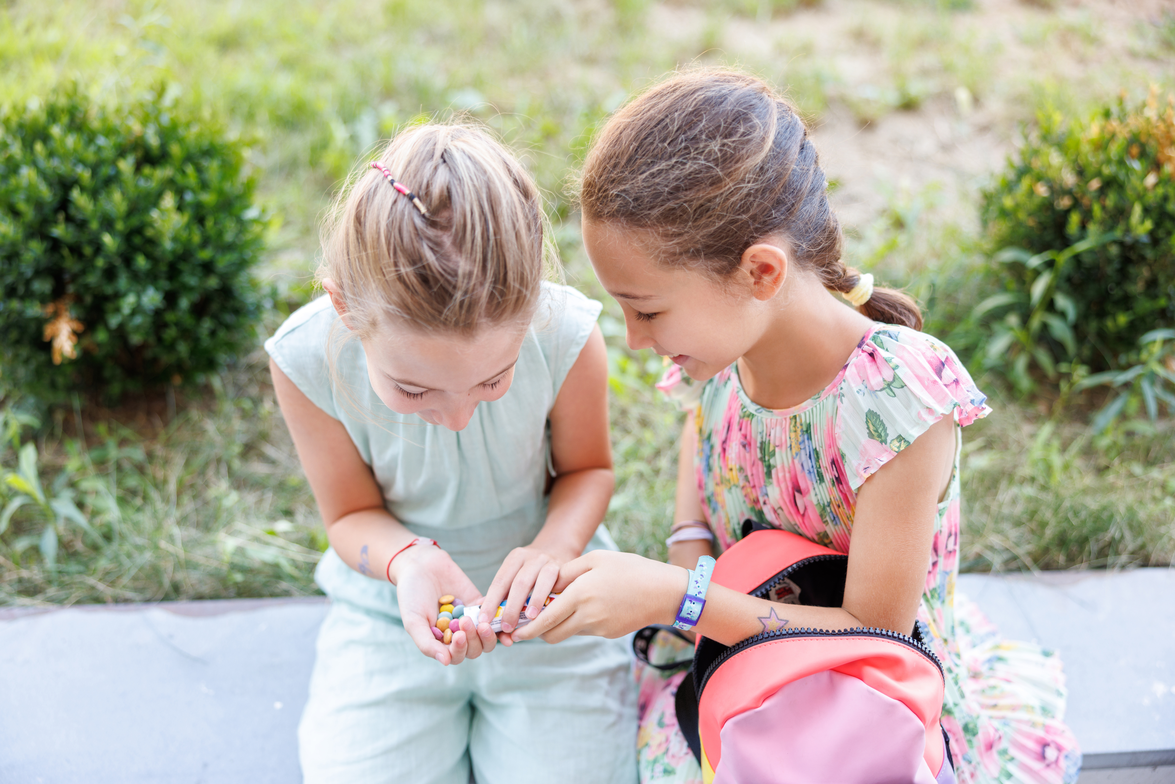 Two young girls examining small objects in their hands while sitting on a sidewalk with bushes behind them. The girl on the right holds a pink backpack