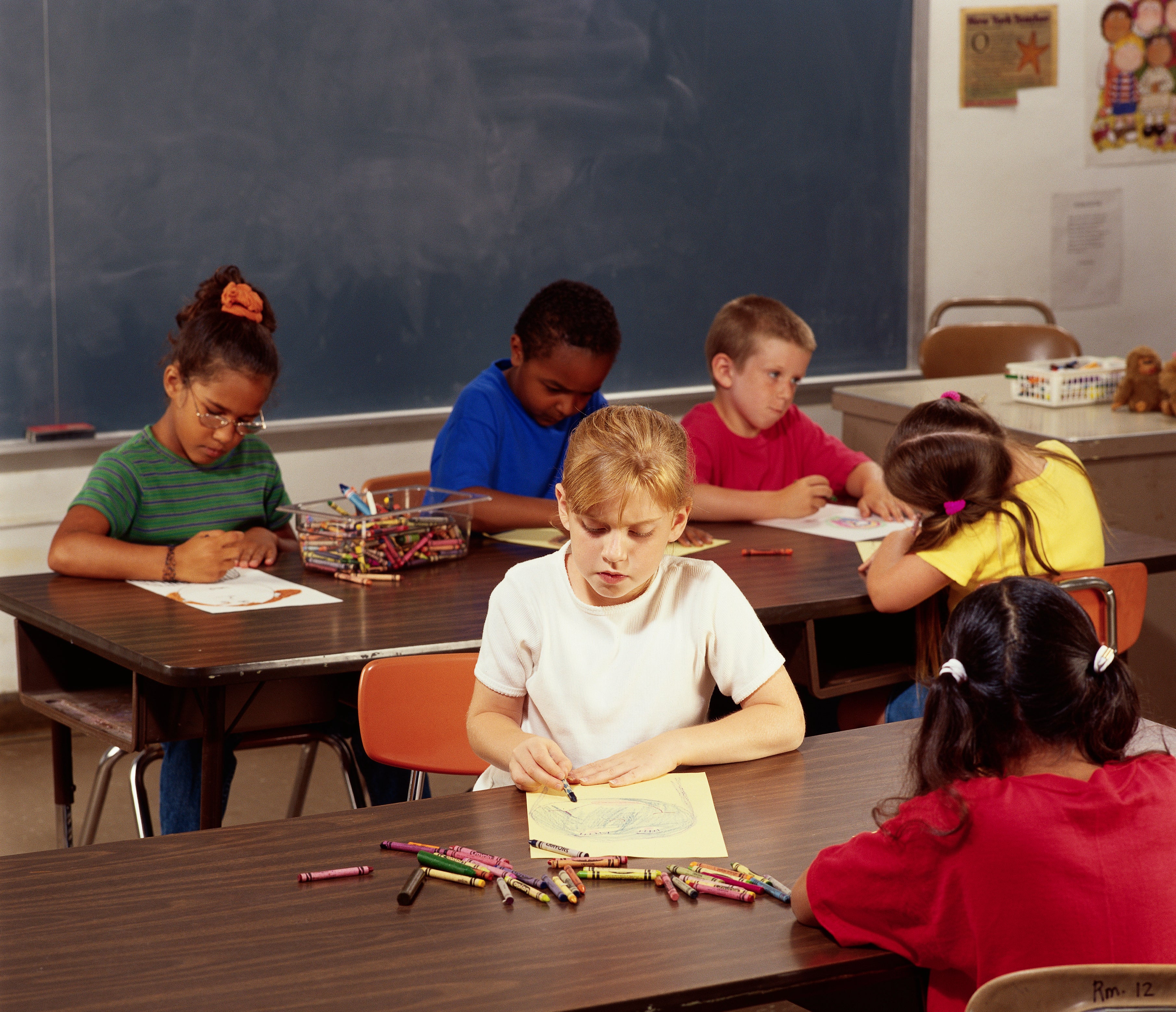 Six young children are seated at desks in a classroom, drawing and coloring with crayons on paper