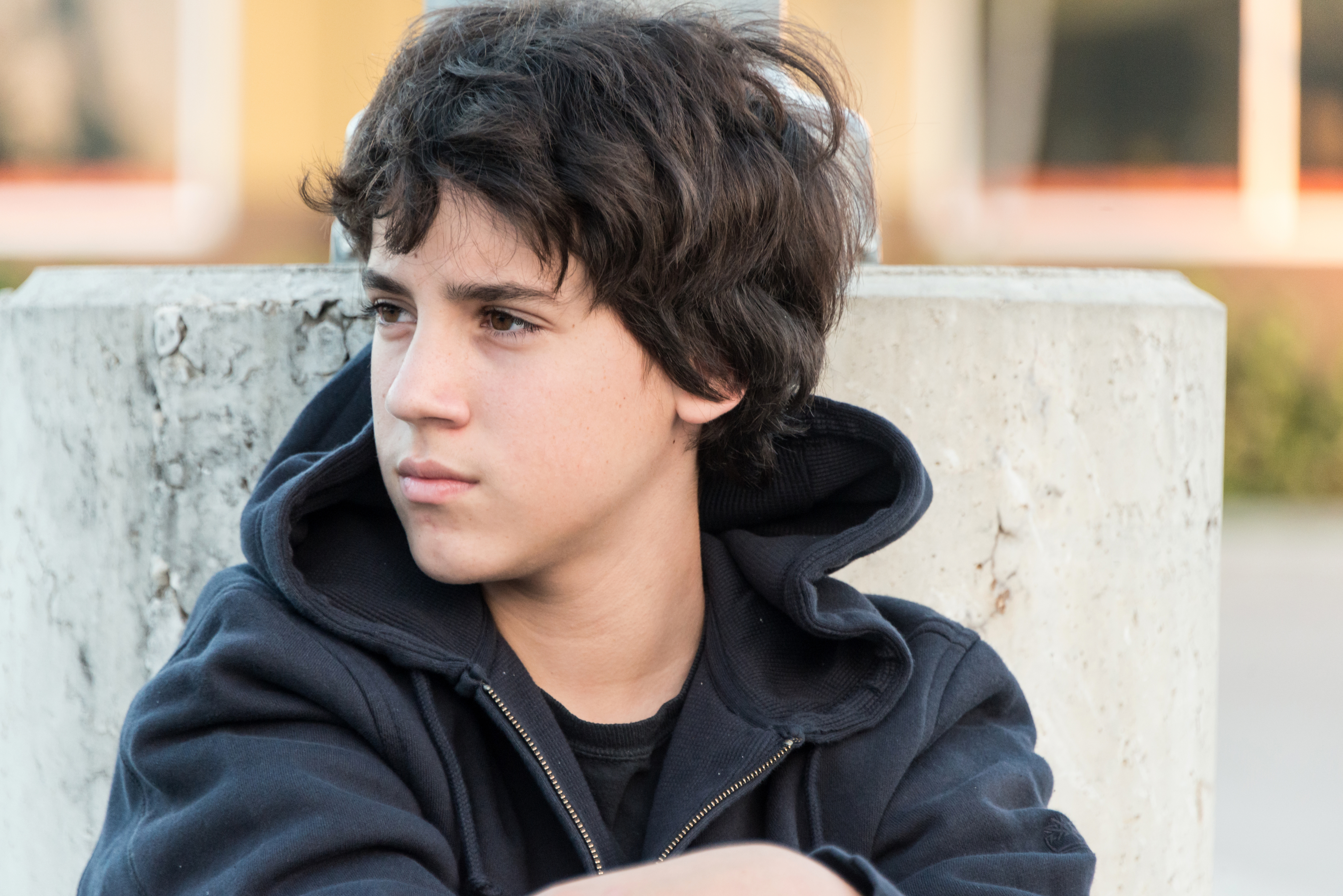 A young person with short wavy hair and a thoughtful expression, wearing a hoodie, sits leaning against a concrete structure outdoors