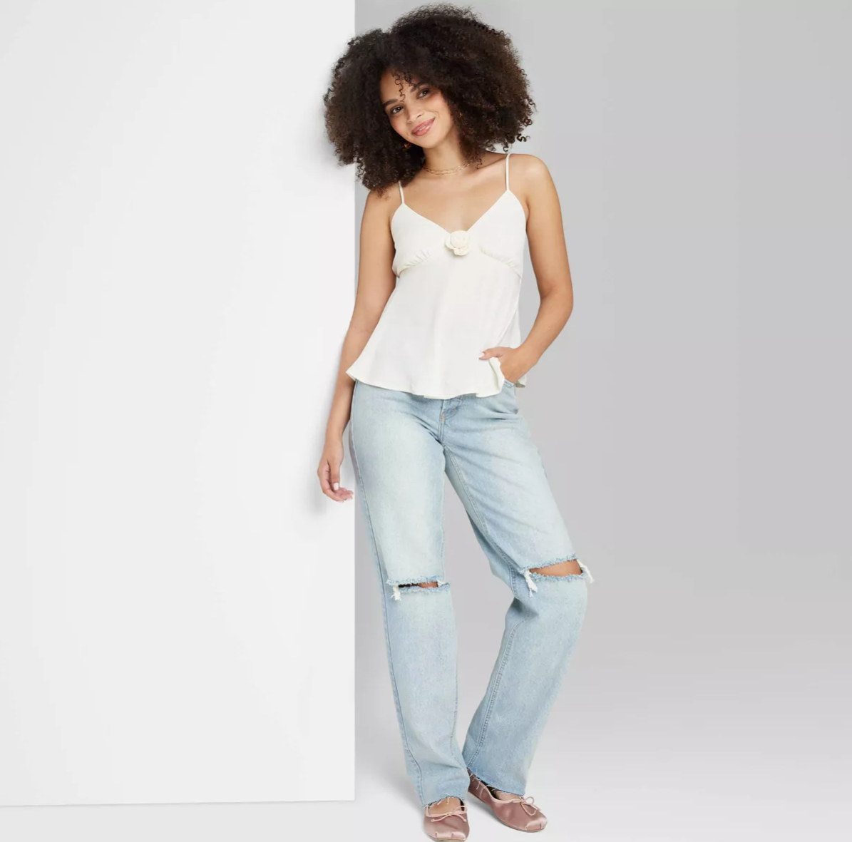 Person with curly hair wearing a white tank top, light blue ripped jeans, and pink shoes, leaning against a white wall