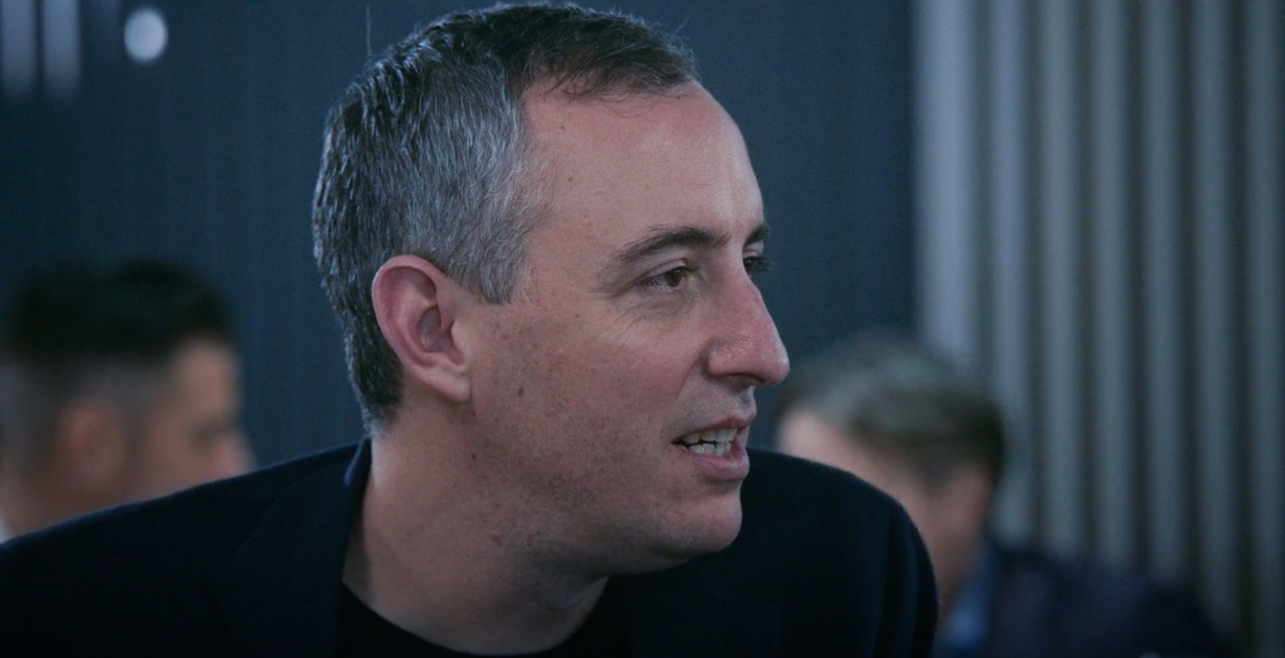 A man is seen in a candid moment, looking to his left, wearing a dark blazer and a black shirt. Background shows blurred people and vertical lines. Names unknown