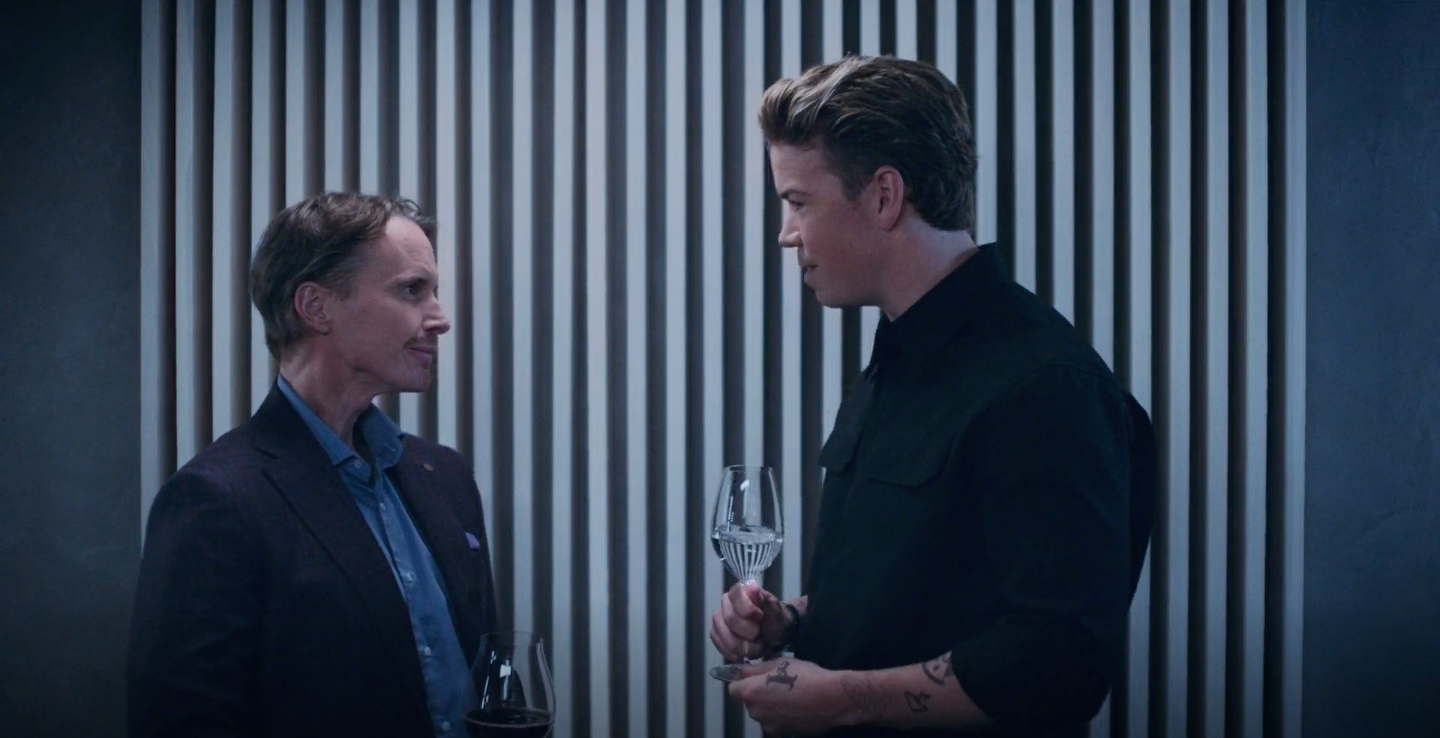 Two unidentified men in formal attire have a discussion while holding wine glasses. There is a striped backdrop behind them