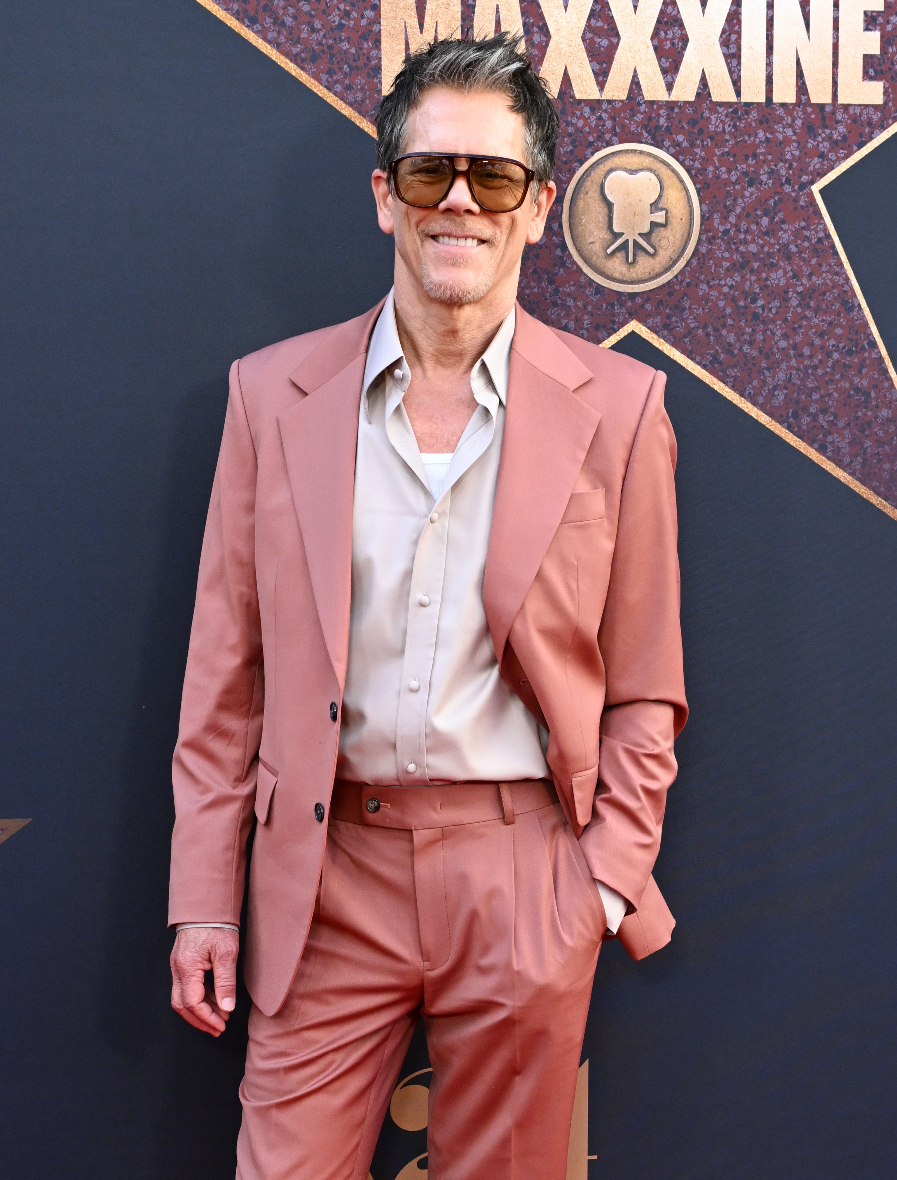 Kevin Bacon stands on the red carpet wearing a salmon pink suit and sunglasses at the premiere of "MaXXXine"