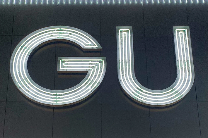 The image displays a neon sign with the letters &ldquo;GU&rdquo; in a modern design, mounted on a wall. The sign features clear, bright white outlines