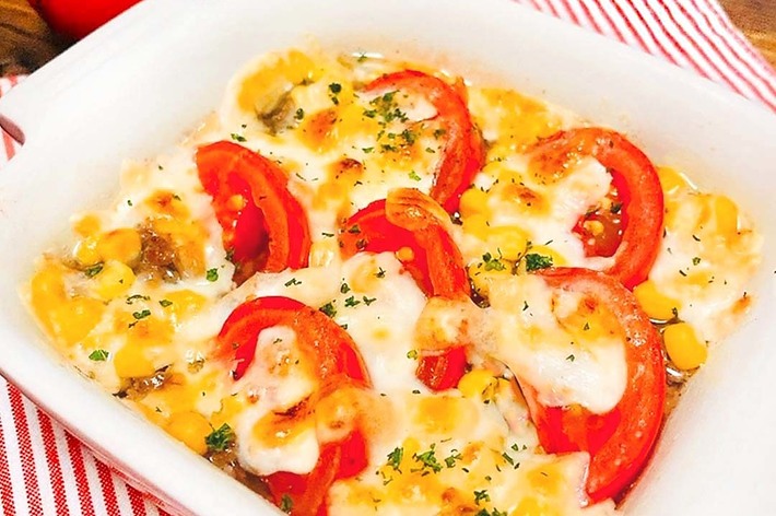 白いベーキング皿に入ったトマトとチーズのグラタン。2つのトマトが背景に置かれた木製のテーブル上にある。