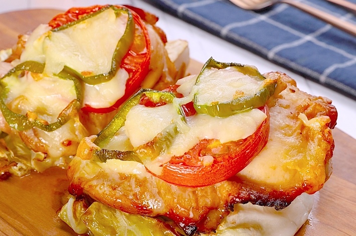 チーズ、トマト、ピーマンがのった肉料理が木のボードに盛られている。背景にはカトラリーが置かれた青い布が見える。