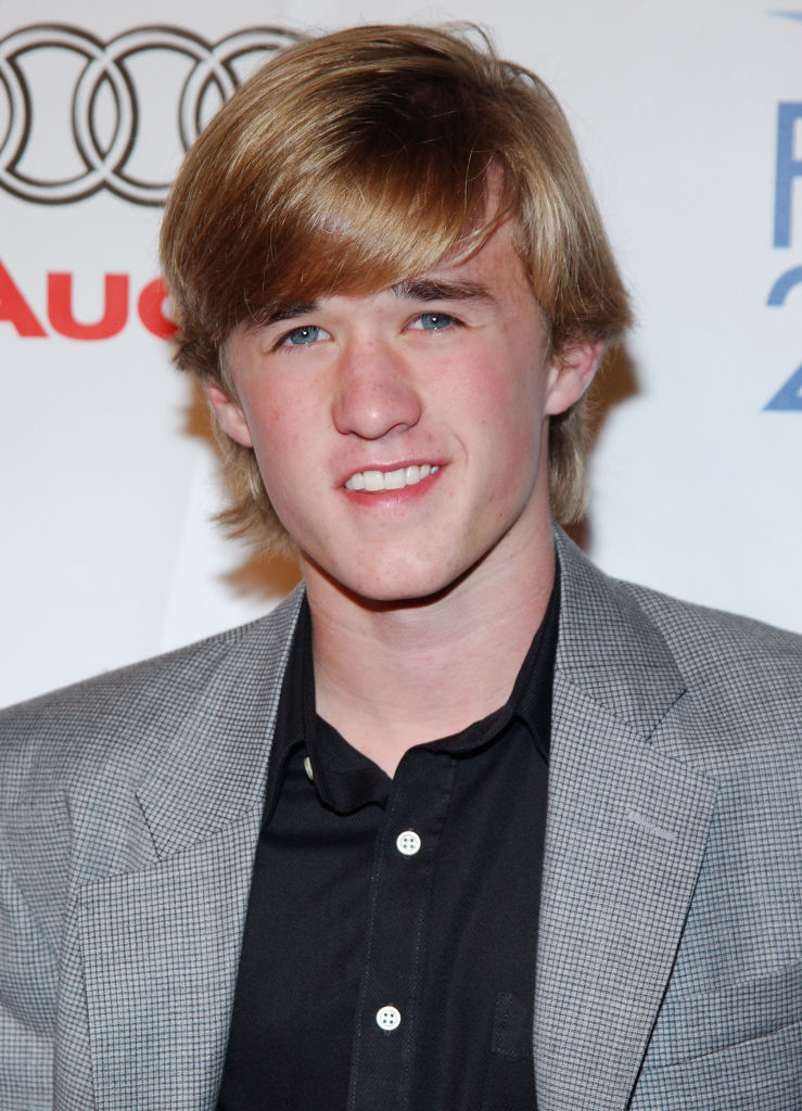 Young man with medium-length blond hair wearing a grey checkered blazer and black shirt, smiling in front of a step-and-repeat banner