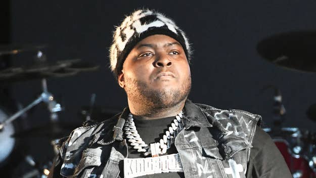 Sean Kingston wears a patterned hat, thick chain necklace, and a sleeveless jacket on stage, looking focused
