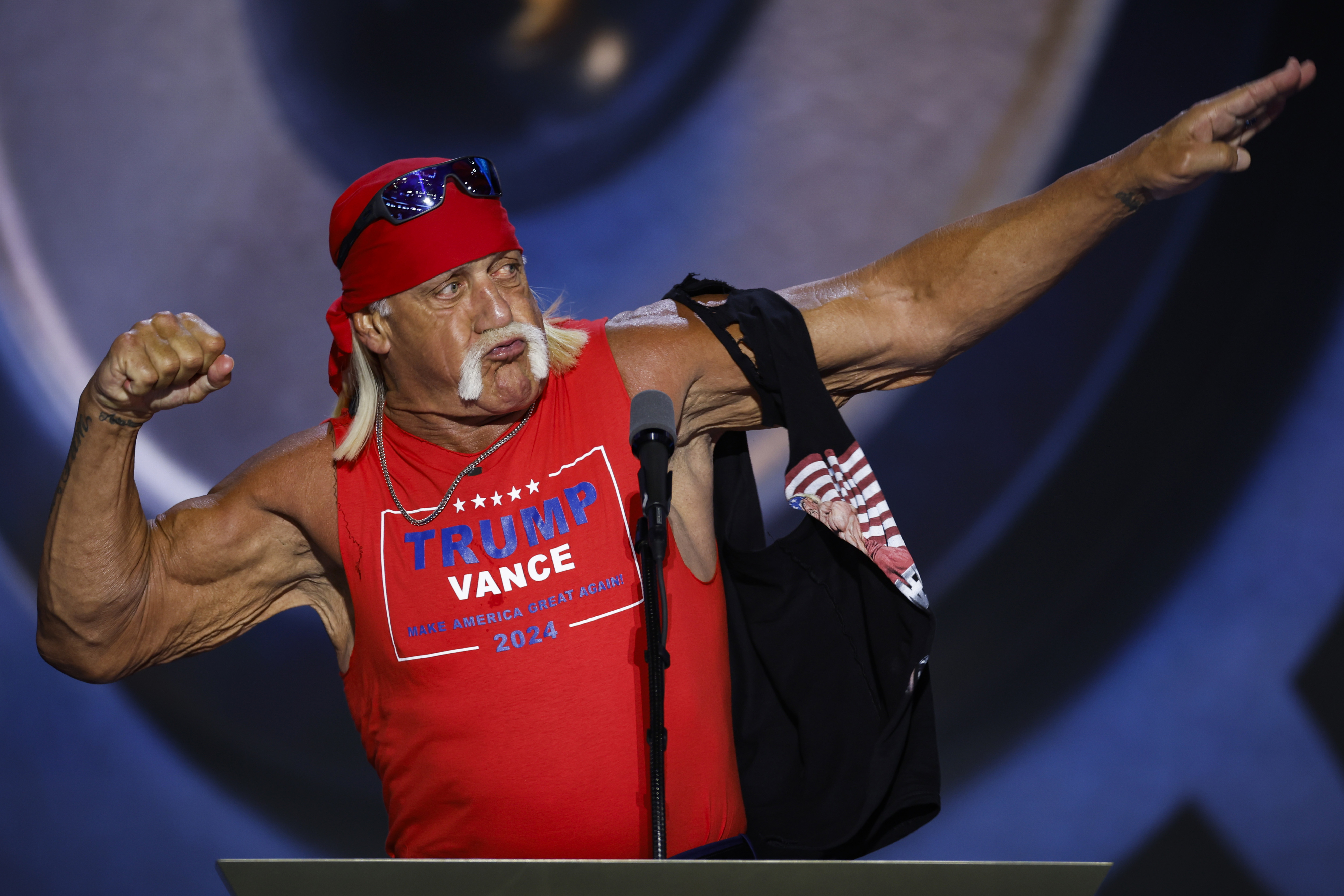 A muscular man wearing a red Trump Vance 2024 tank top and red bandana poses with a flexed arm and a pointed finger gesture at a podium