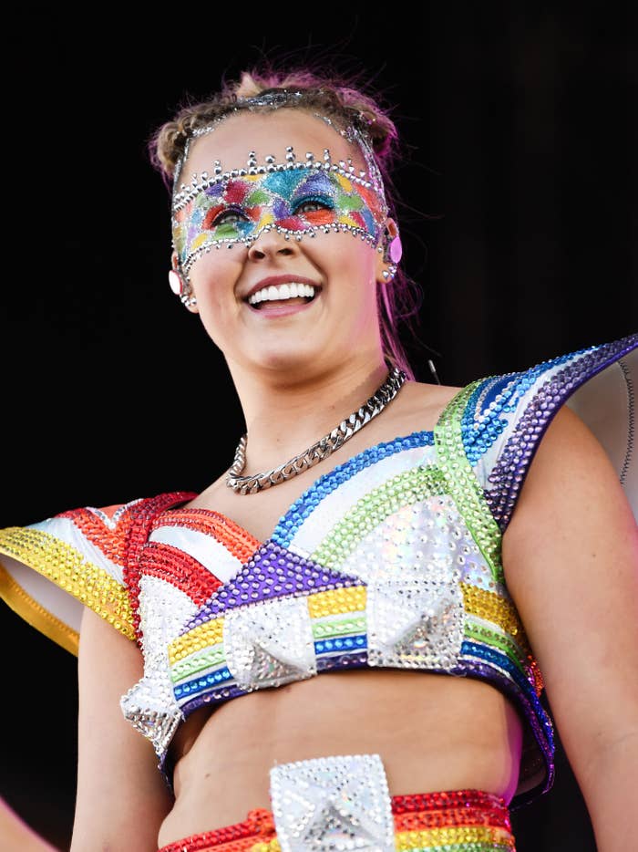 JoJo Siwa wearing a sparkly, colorful outfit with an embellished mask, smiling and performing on stage