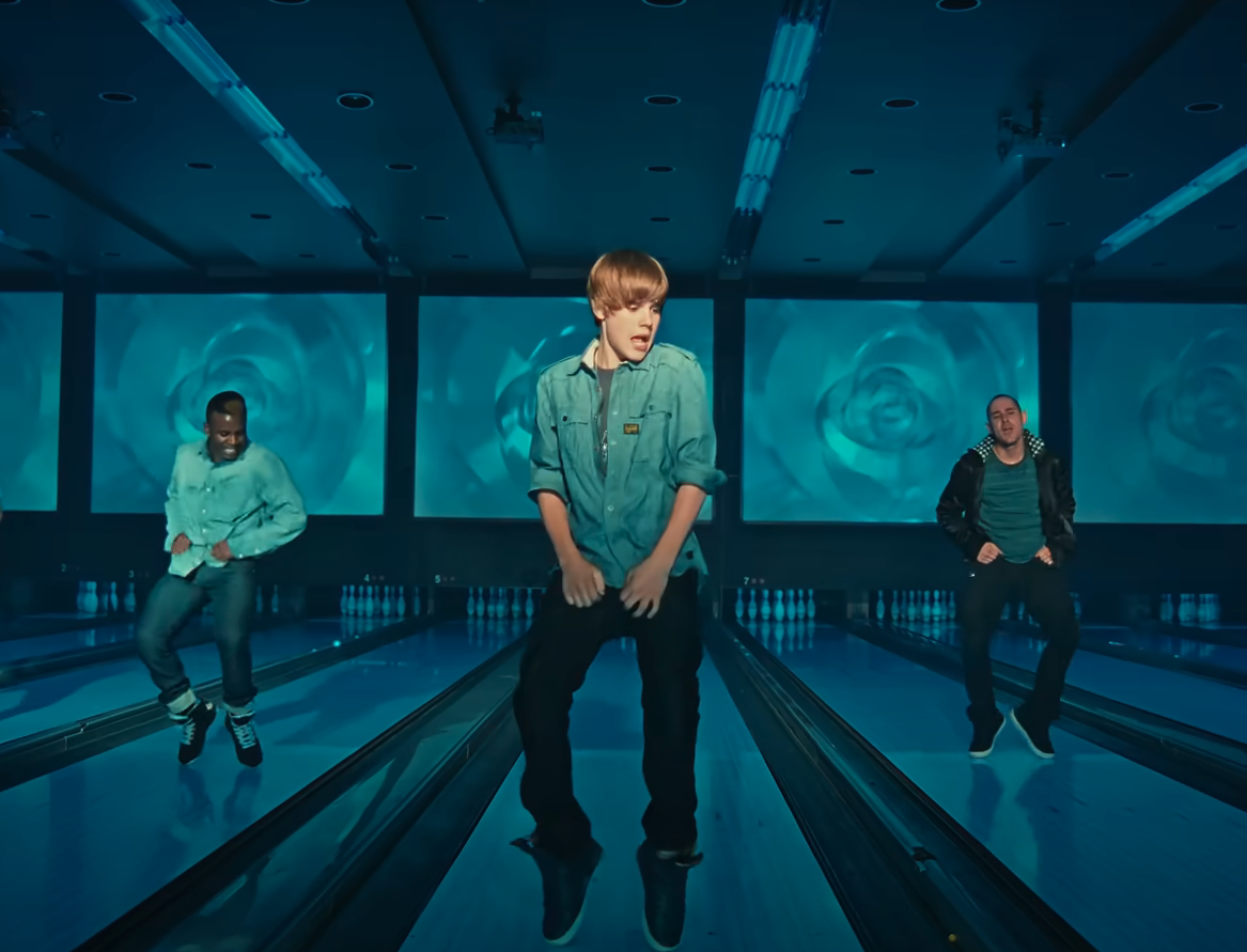 Justin Bieber, Ludacris, and an unidentified person dance in a bowling alley with screens displaying a blue swirl pattern behind them