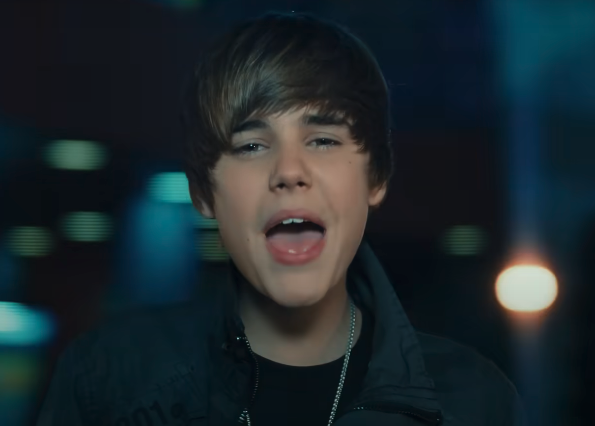 Justin Bieber singing passionately in a music video, wearing a dark jacket and a necklace