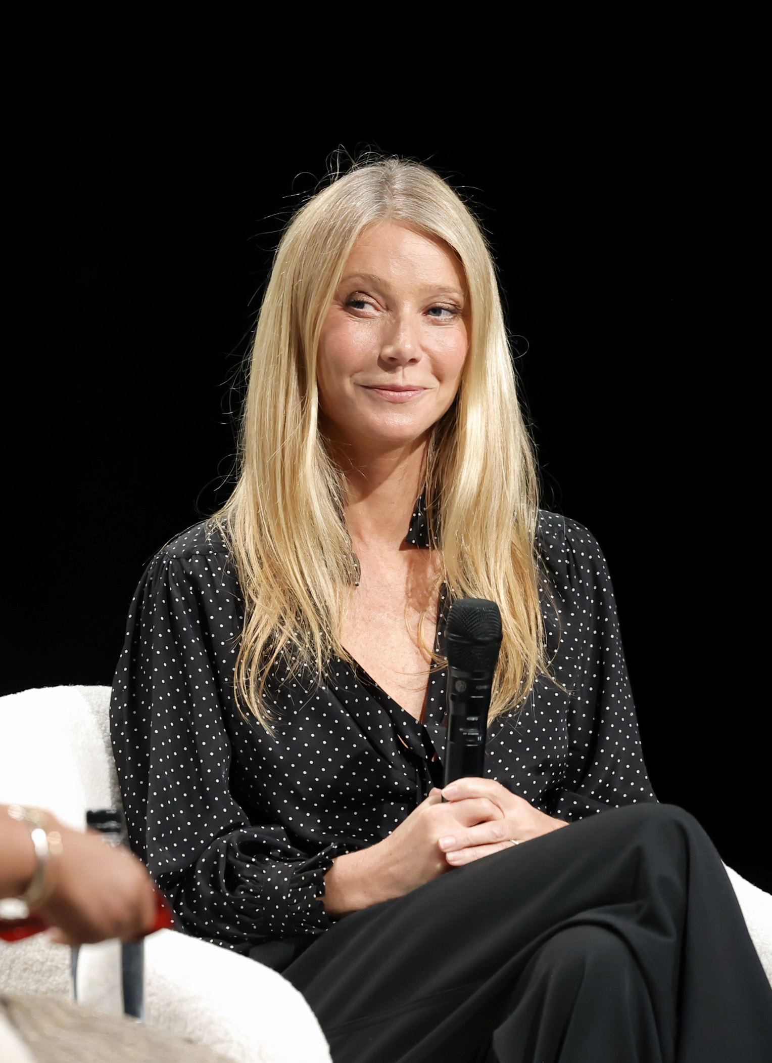 Gwyneth Paltrow speaking into a microphone while sitting on a stage, wearing a black long-sleeve blouse with white polka dots and black pants