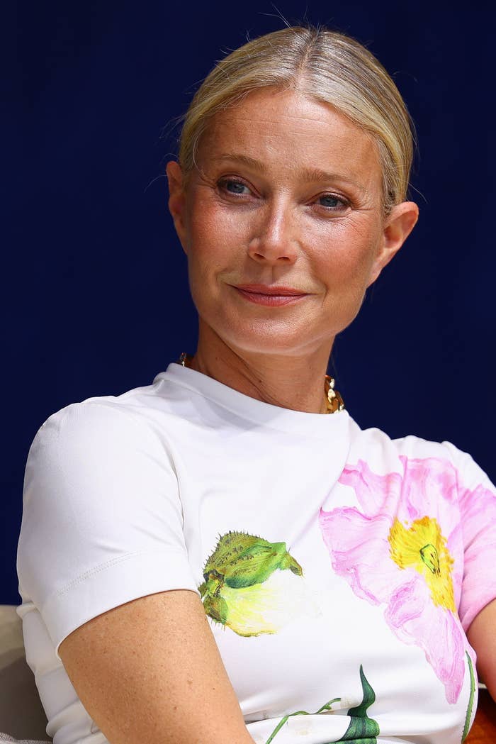 Gwyneth Paltrow wearing a white top with floral prints, smiles slightly, posing against a dark background