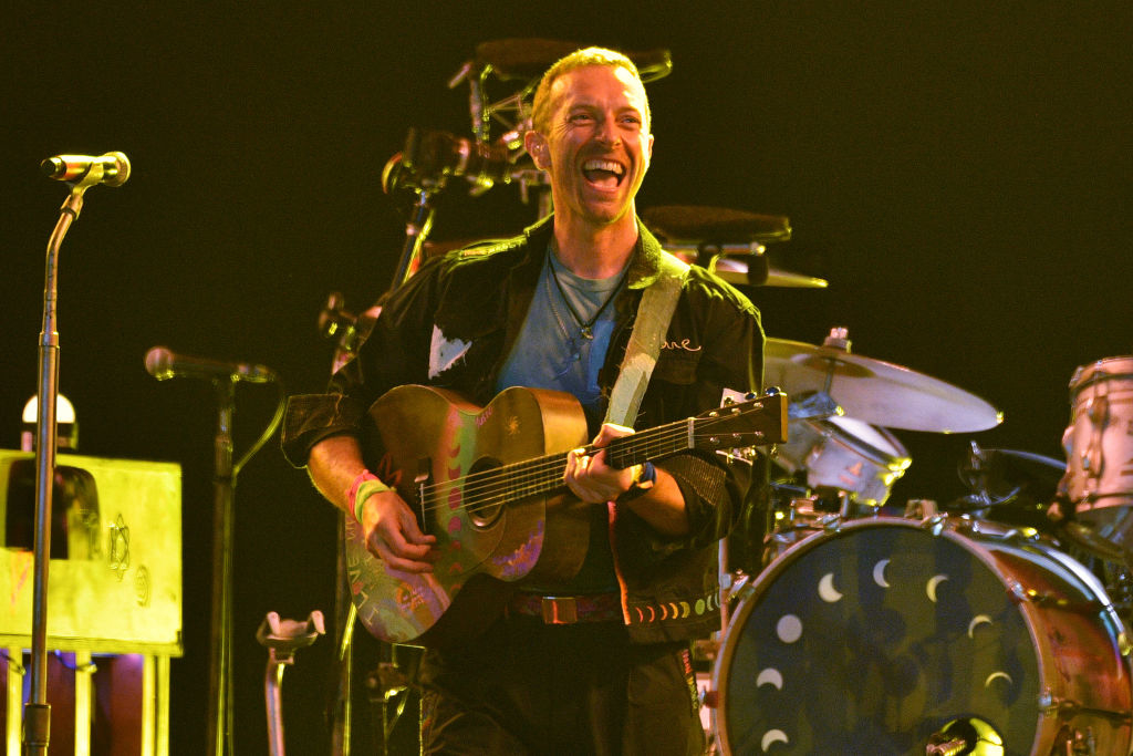Chris Martin, smiling broadly while playing a guitar with colorful designs, performs on stage with drums and other instruments in the background