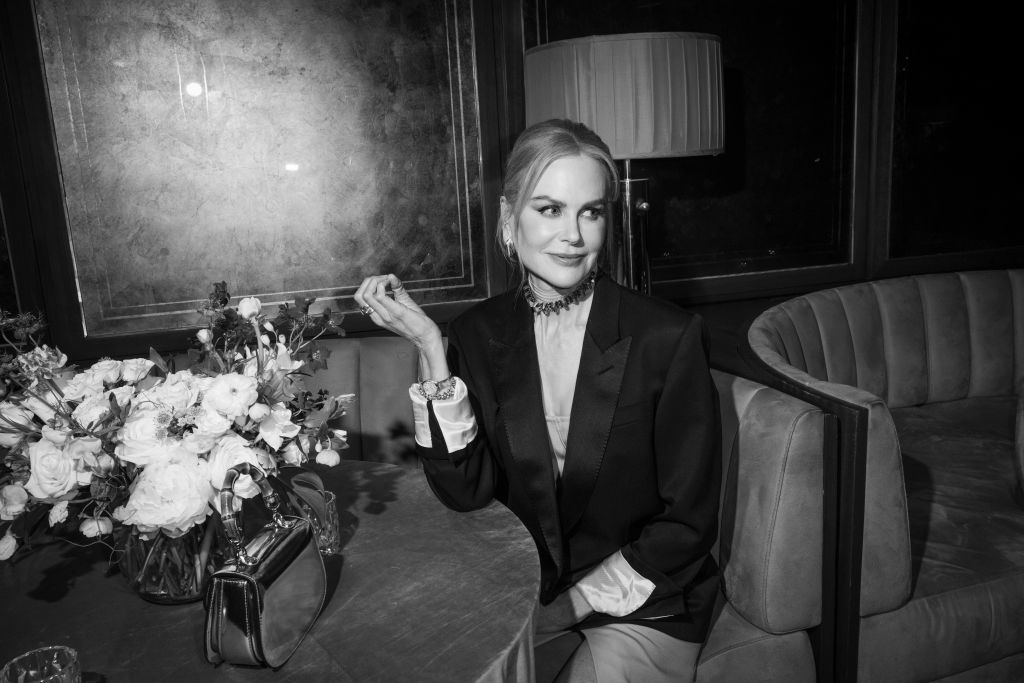 Nicole Kidman sits at a table in a formal setting, wearing an elegant blazer and choker necklace, with a floral arrangement and handbag on the table