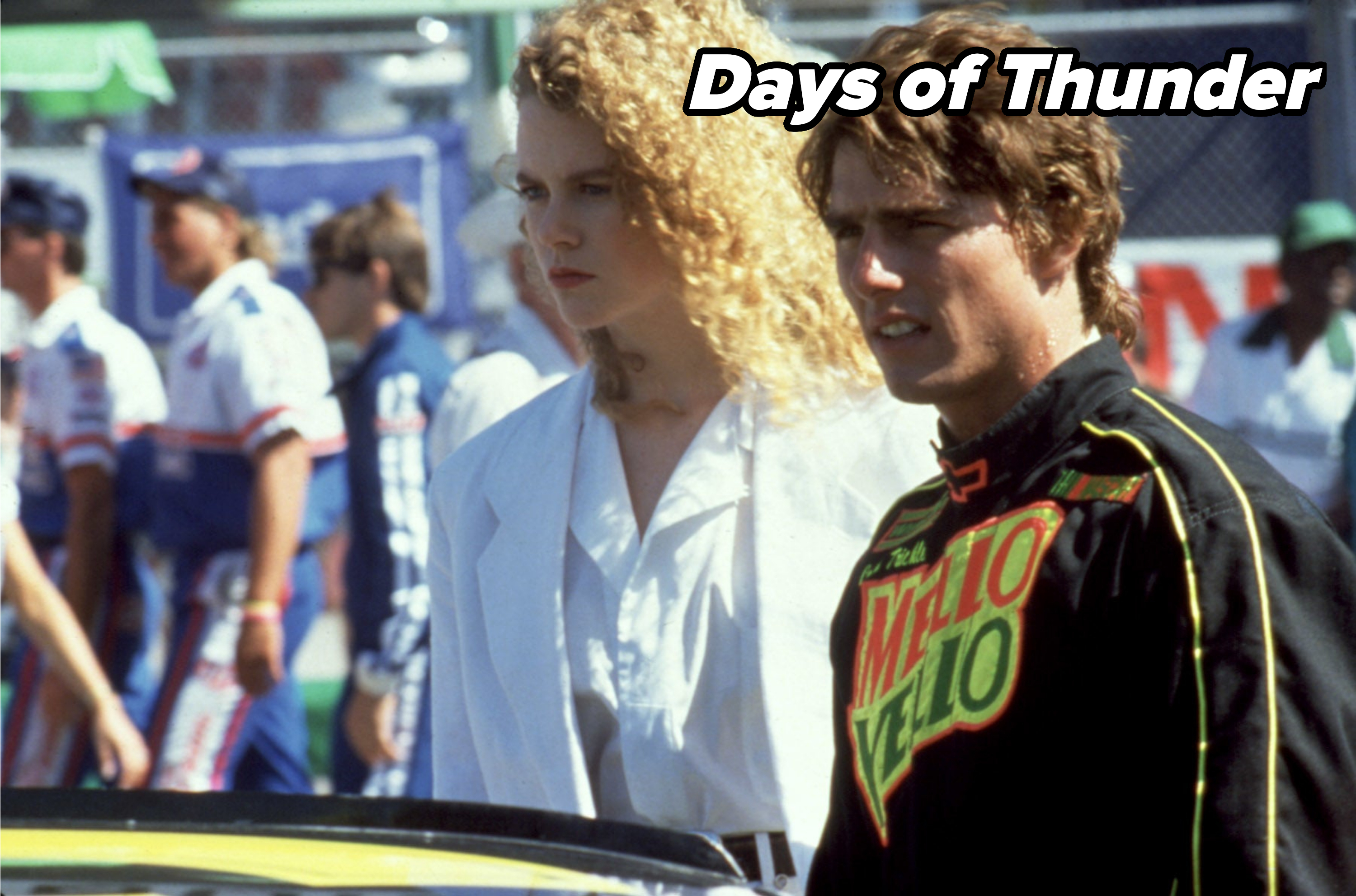 Nicole Kidman and Tom Cruise stand together at a racing event. Nicole wears a stylish white outfit, while Tom wears a racing suit with "Mello Yello" branding