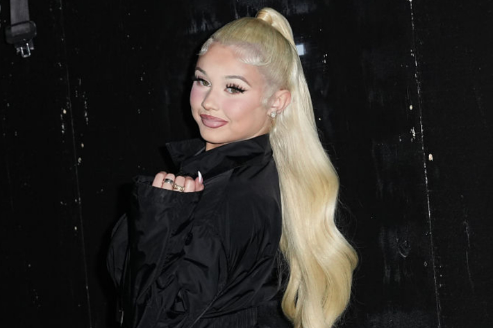 A person with long, blonde hair in a high ponytail poses in a black outfit against a dark background