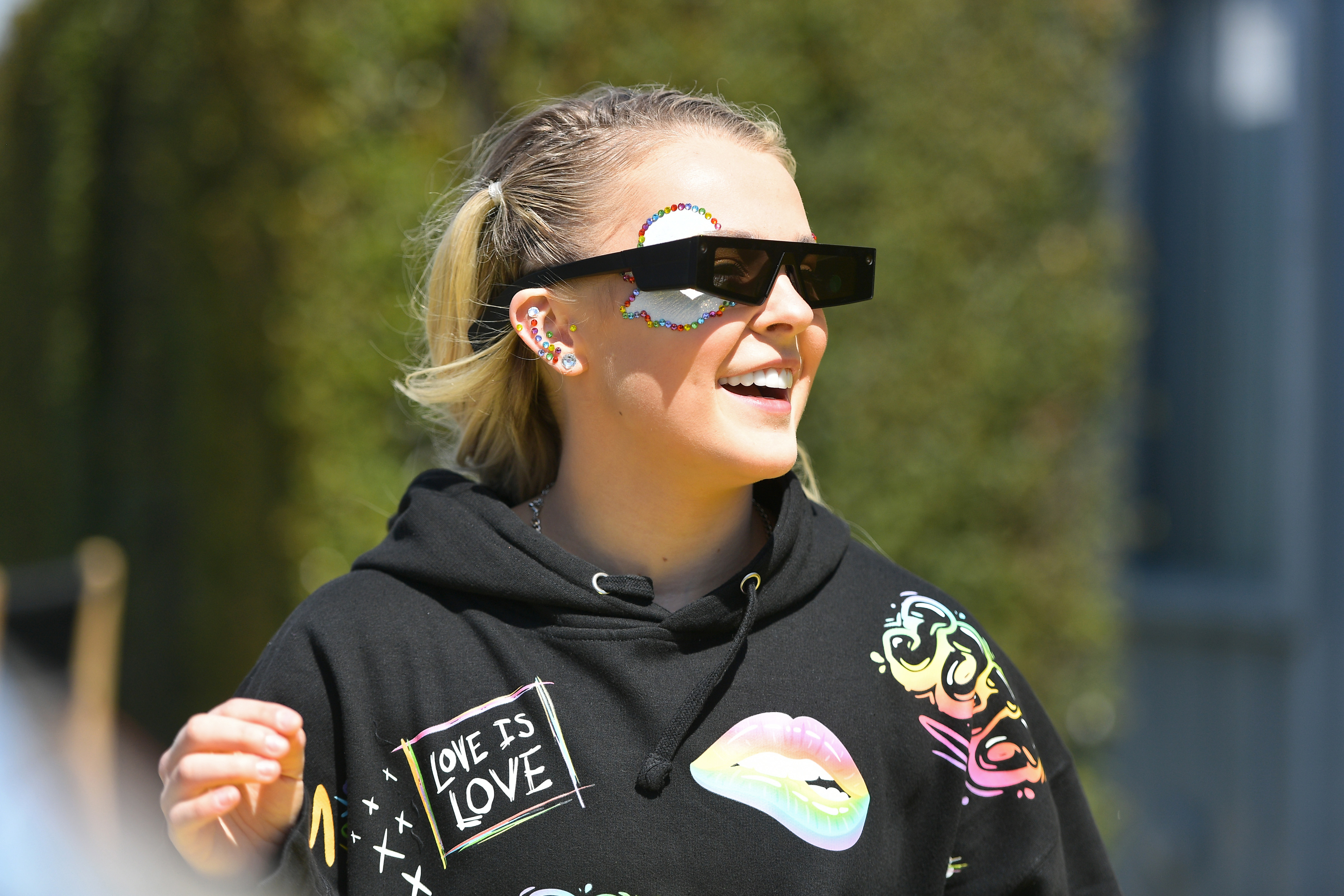 JoJo Siwa wearing oversized sunglasses and a black hoodie with colorful patches and "Love is Love" text, smiles outdoors