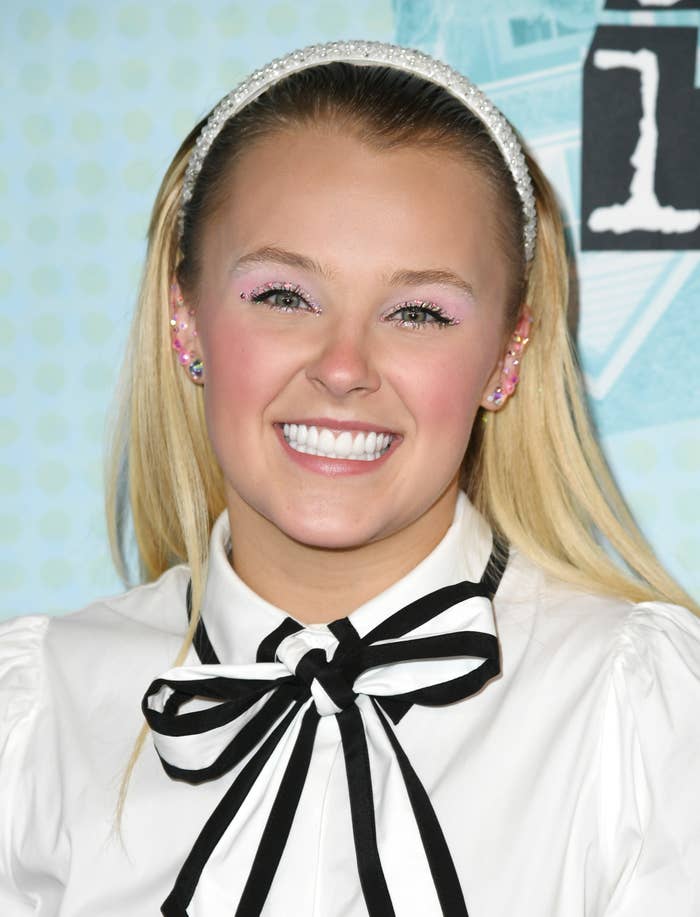 JoJo Siwa smiles at an event, wearing a white blouse with a large black-and-white bow tie and a sparkly headband