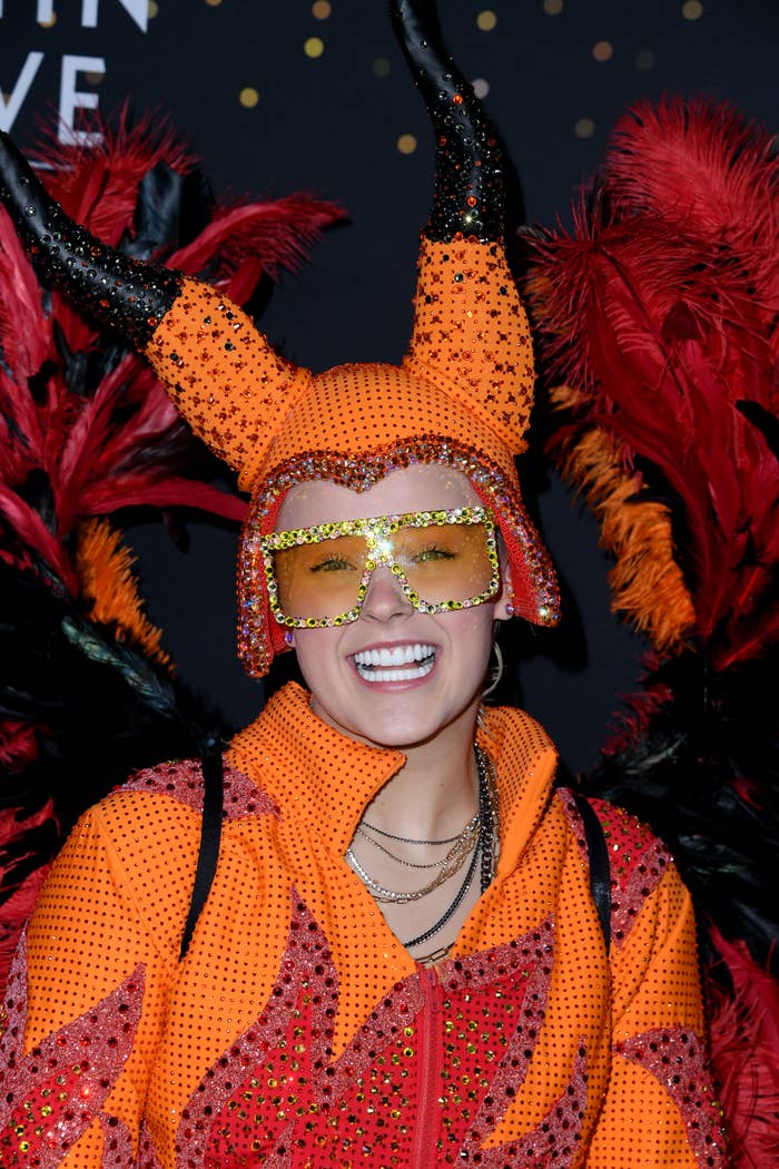 JoJo Siwa smiles on the red carpet in an orange, sequin-studded devil costume with large horns and red feathered wings
