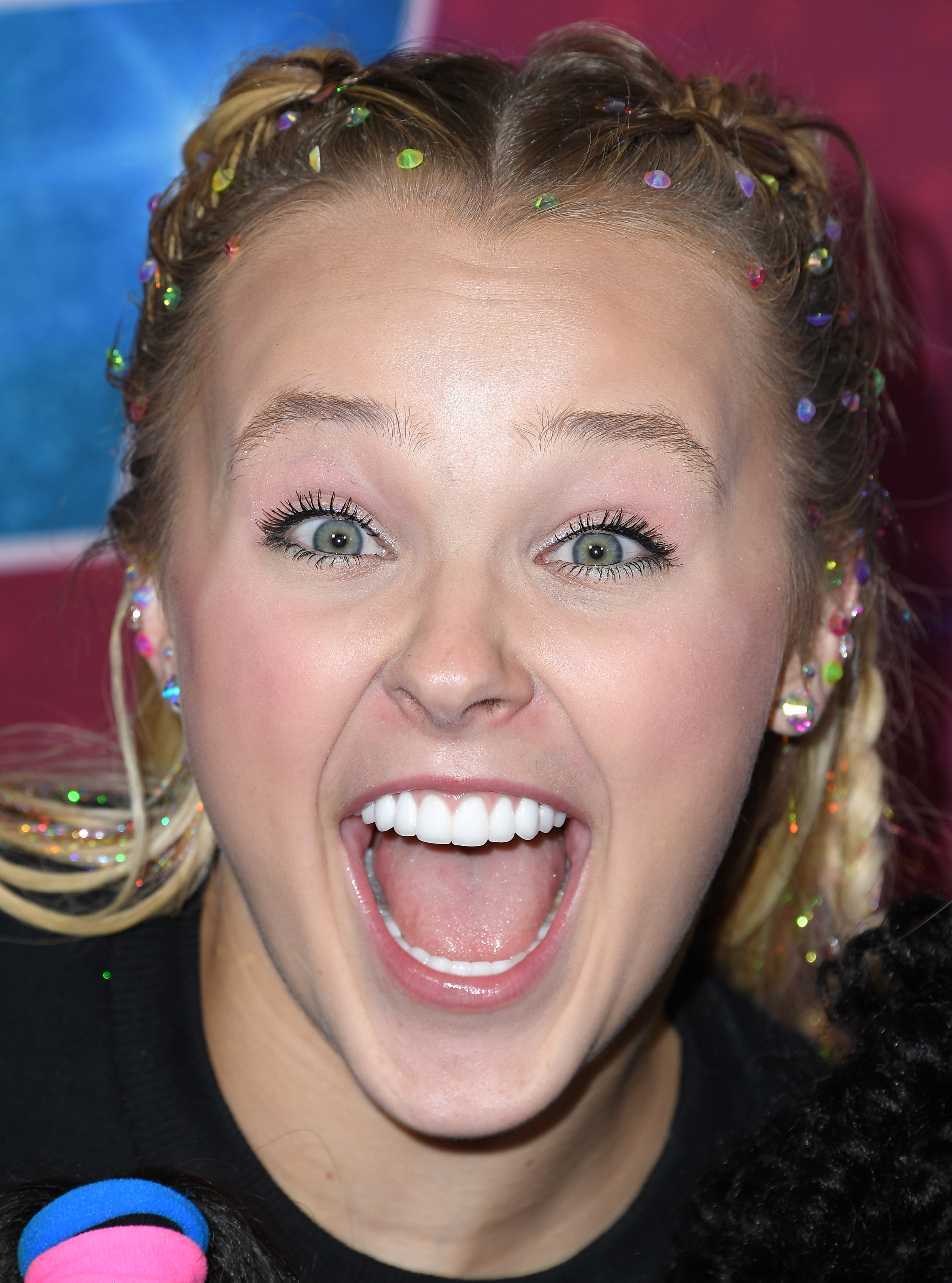 JoJo Siwa smiling on the red carpet, with hair styled in braids and decorated with colorful gems