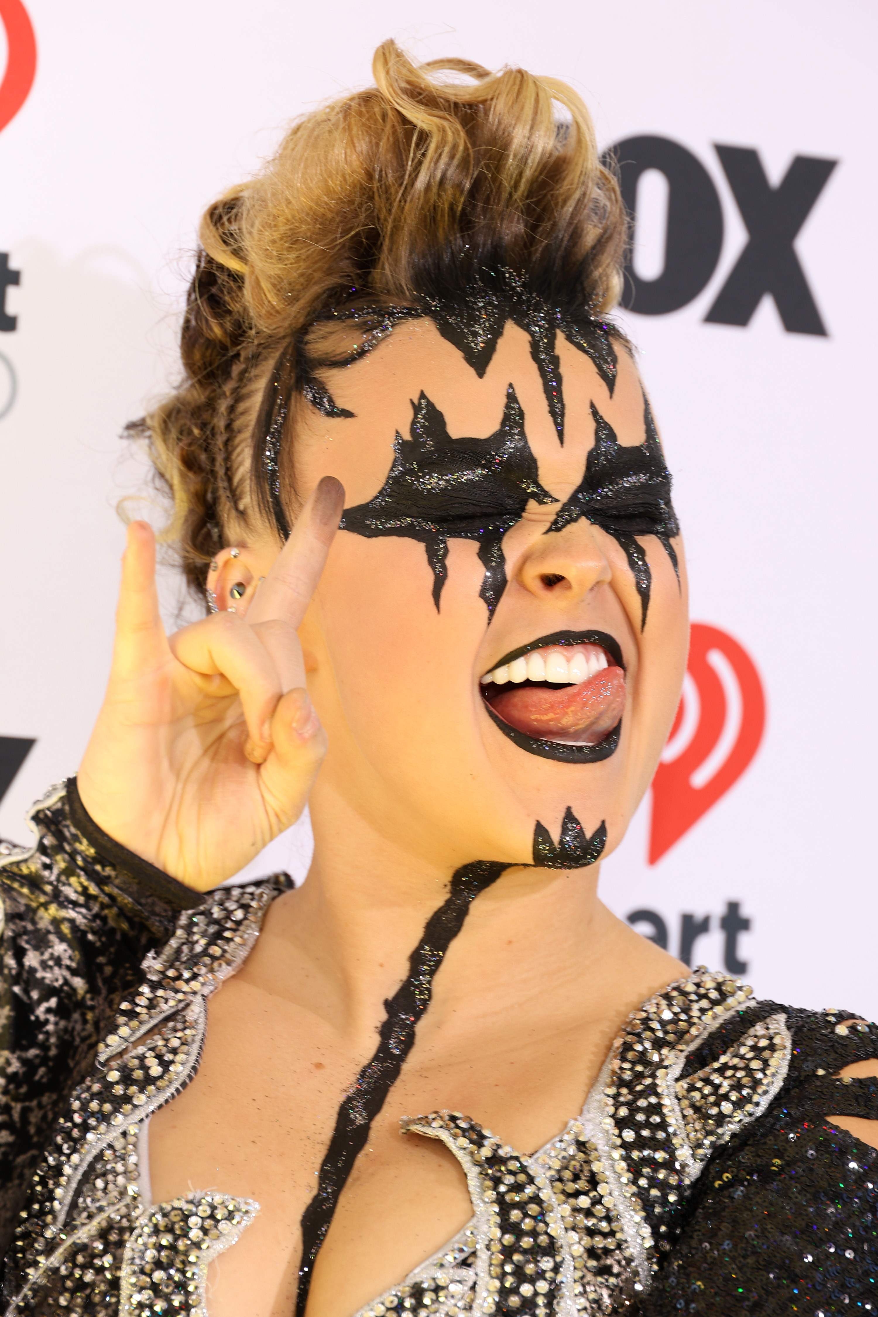 Person dressed in an elaborate, rock-inspired outfit with black face paint and decorated clothing, making a rock hand gesture and sticking out their tongue