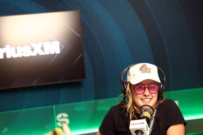 JoJo Siwa in a SiriusXM studio, wearing a white cap, large pink sunglasses, and headphones, speaking into a microphone
