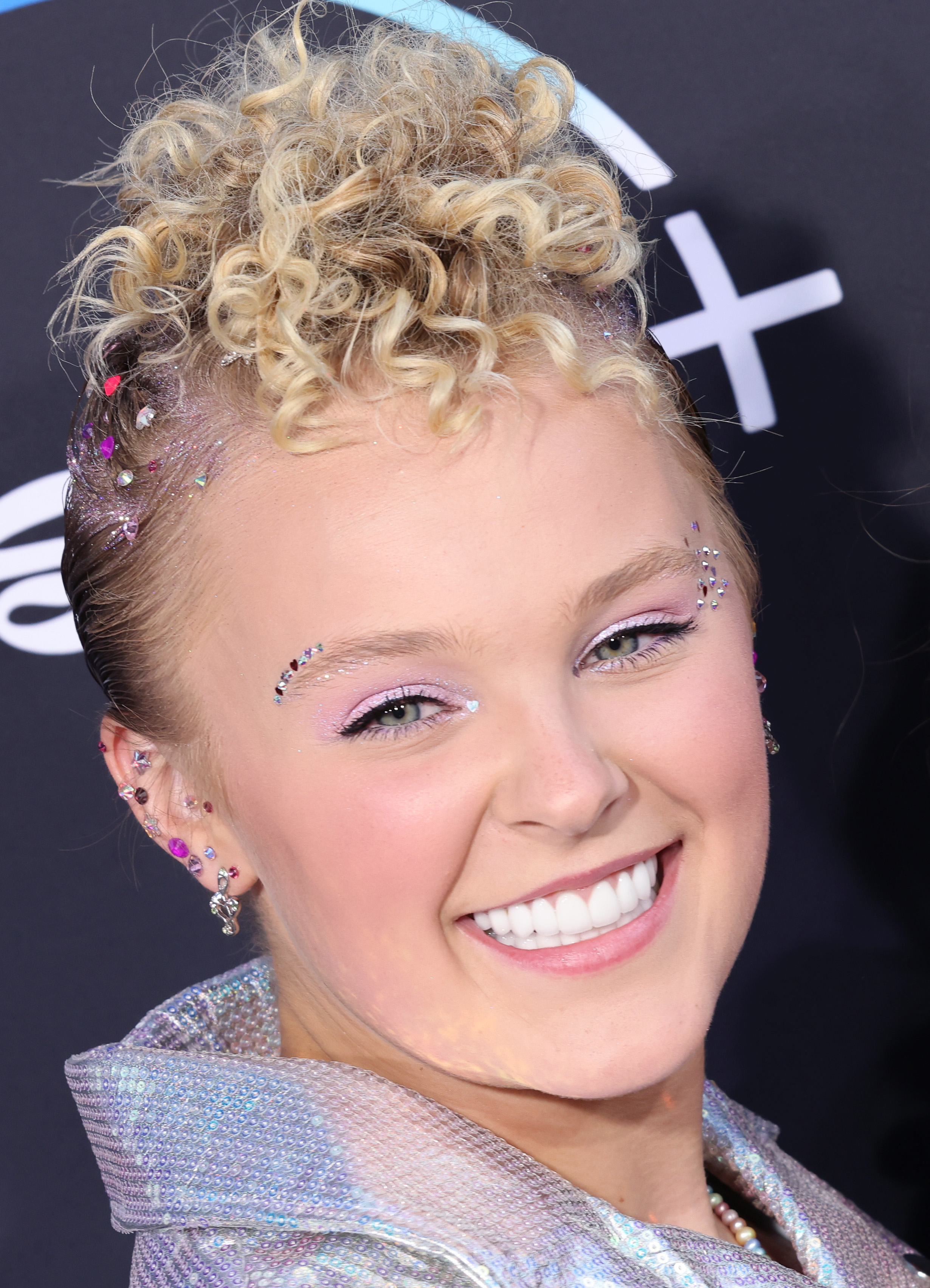 JoJo Siwa smiles on the red carpet with a curly updo and glittery makeup, wearing a shiny, metallic outfit