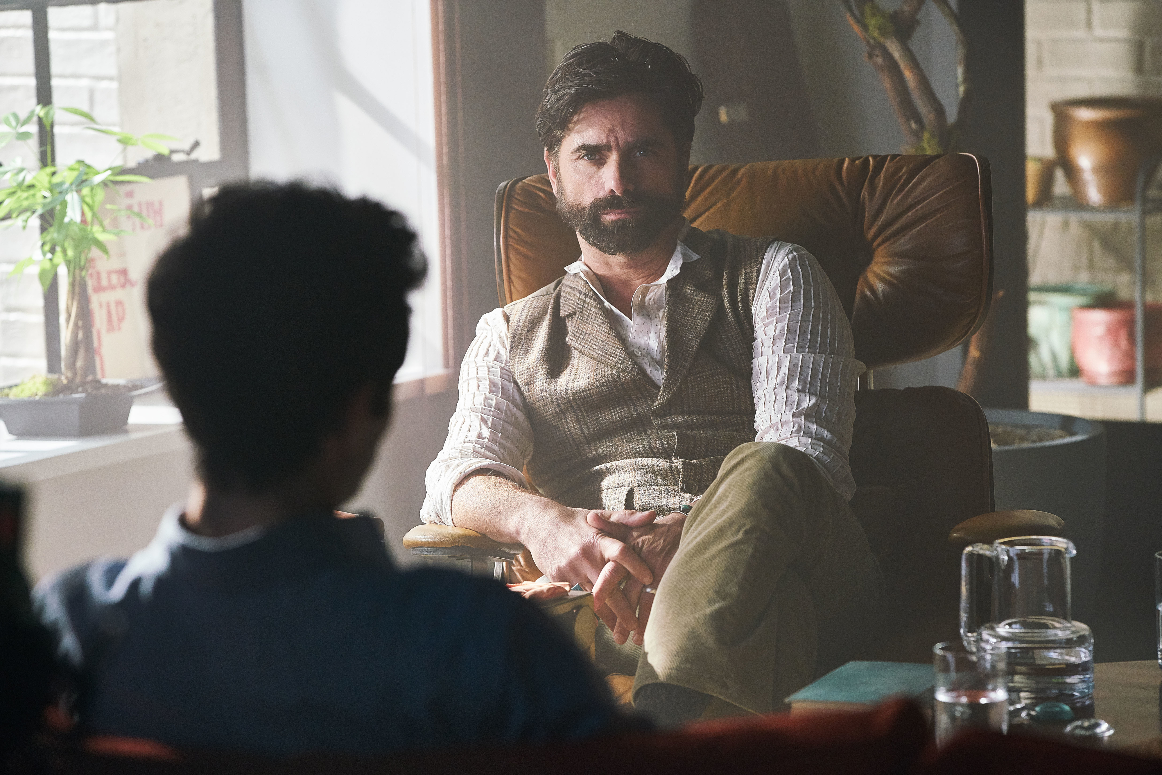 John Stamos dressed casually in a waistcoat, shirt, and trousers, sitting in a mid-century modern chair, conversing with a person whose back is to the camera